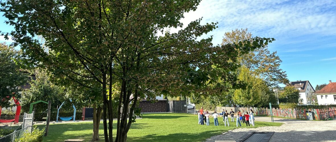 Kinder halten sich im Kreis auf einer Spielwiese vor Bäumen und Häusern.