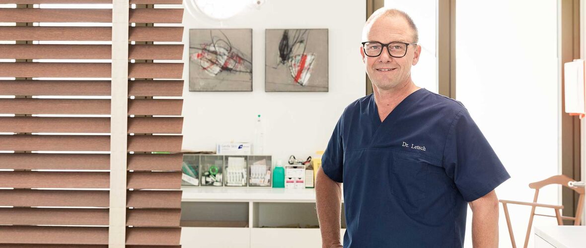 Dr. Letsch lächelnd in blauer Arztkleidung vor medizinischer Einrichtung